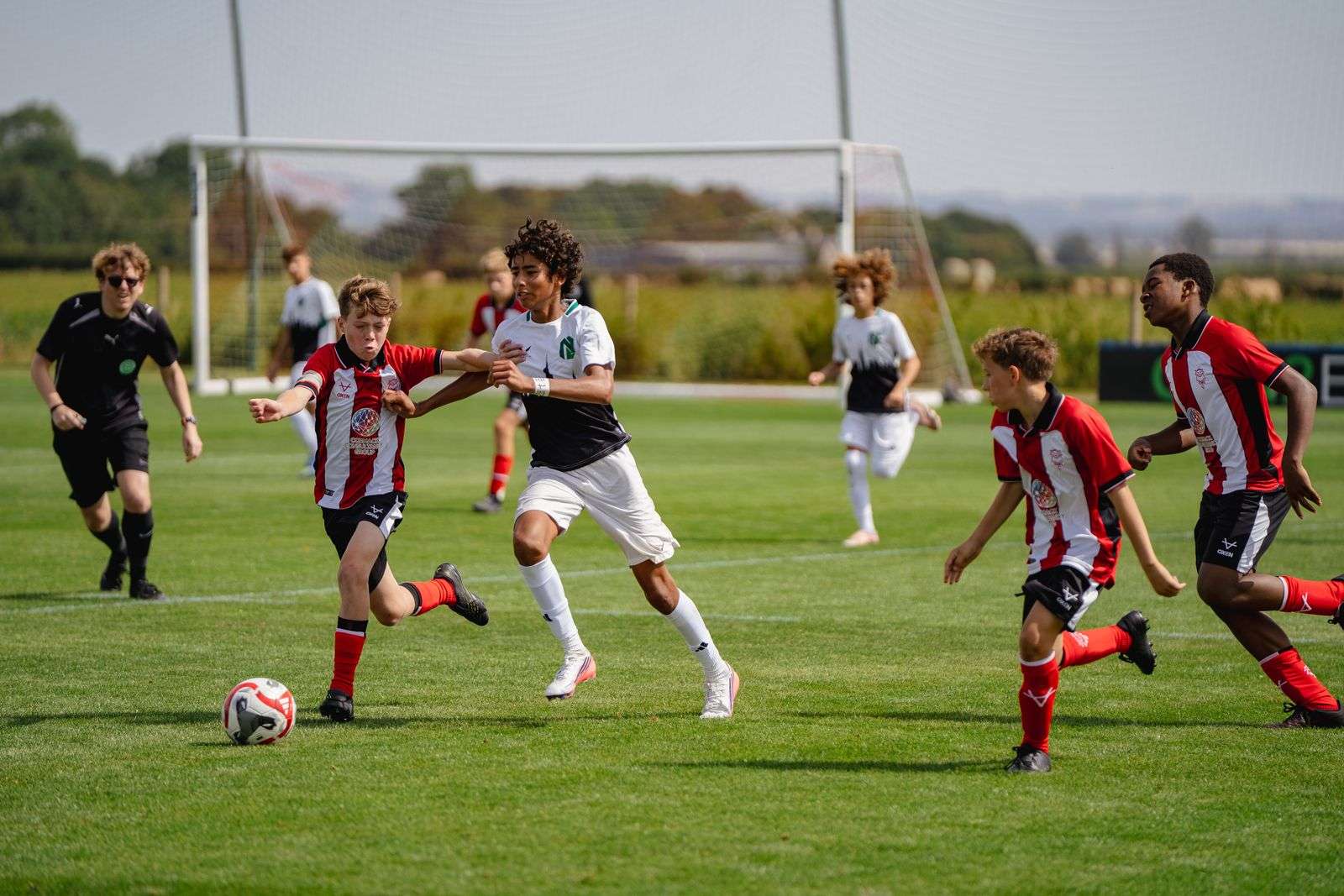 Player in white runs at defenders in red and white stripes as a referee watches, football trials in England