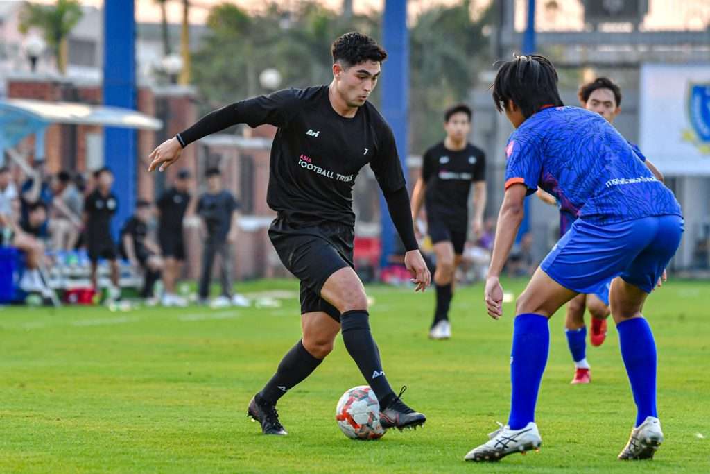 Player in a black kit dribbling past a player in blue at an Asia football trial