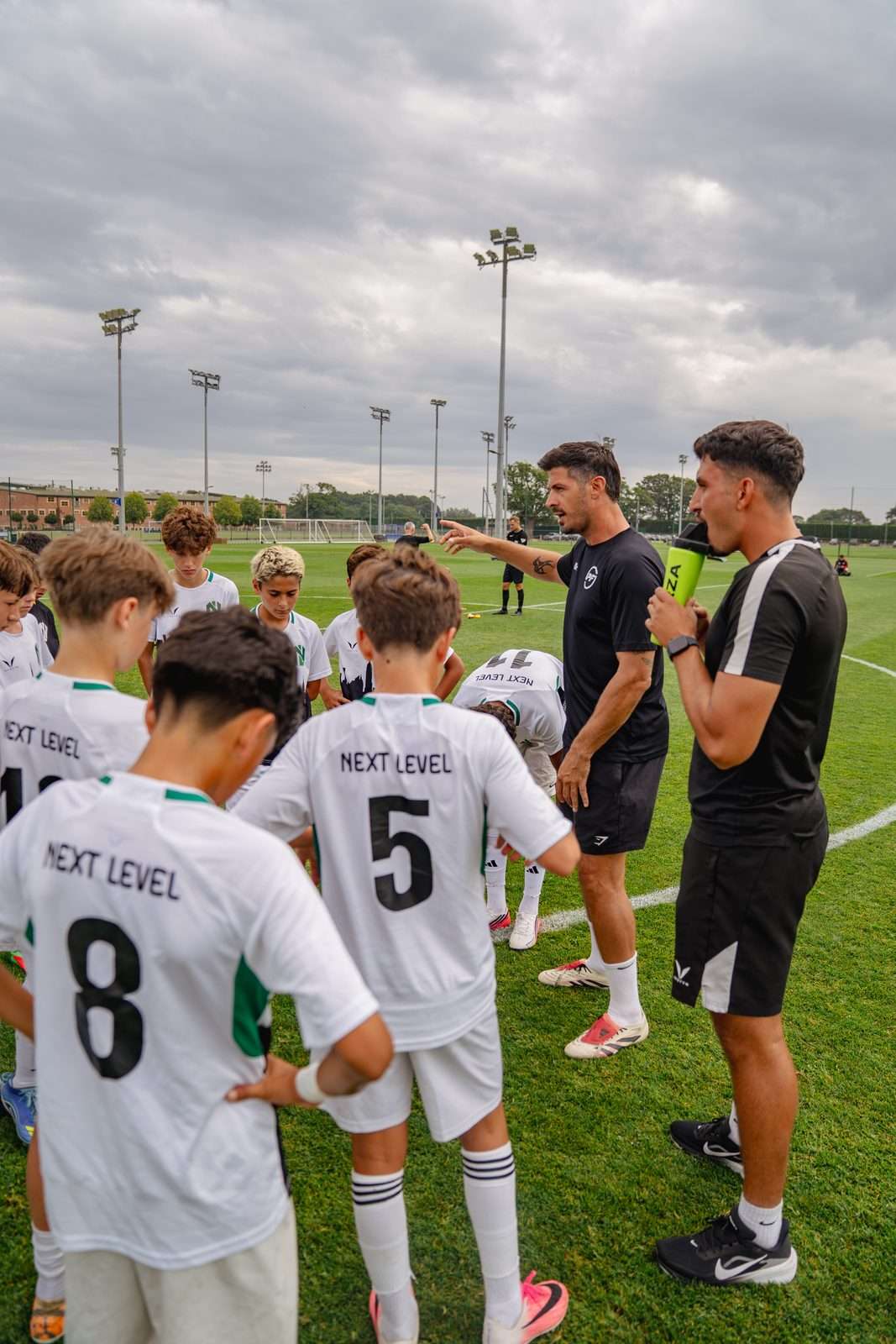Coaches in black give a team talk to players in white Next Level shirts at a UK football trial