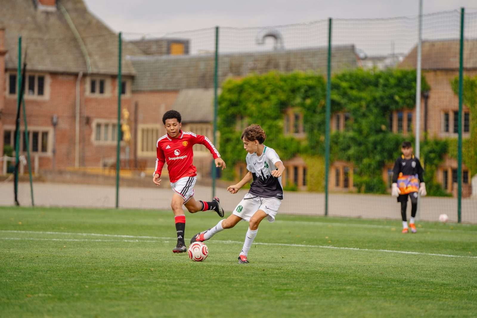 Player in white dribbles towards a defender in red beside brick buildings at a UK football trial